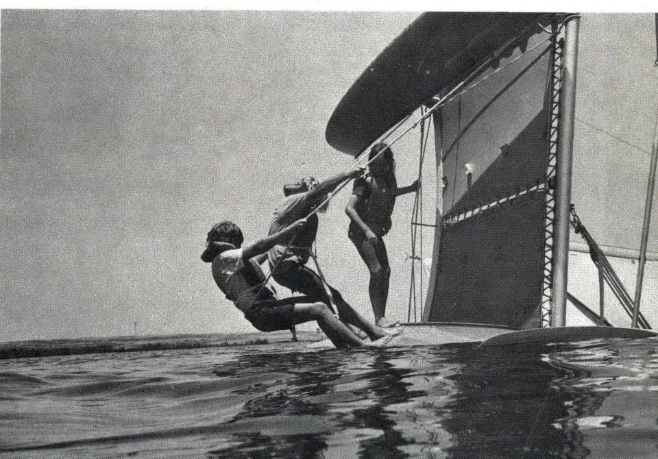 Black and white photo of three children leaning back in a loop of righting line from the lower hull of a capsized Hobie 16 as the mast begins to lift
