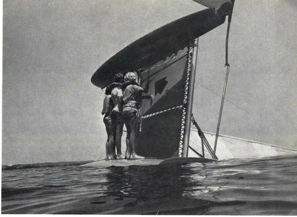 Black and white photo of three children standing on the lower hull of a capsized Hobie 16, one pulling the righting line down from under the trampoline